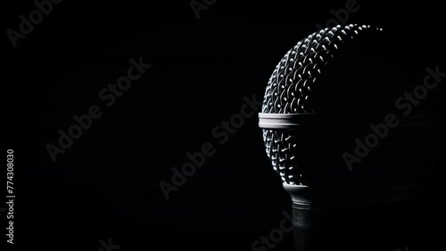 The vocal handheld microphone slowly rotates in backlight on black background. Close-up chrome grid on dynamic mic surface. Copyspace. Concept recording studio, voice, podcast, karaoke, audiobook photo