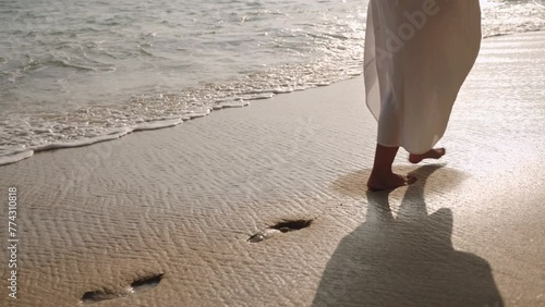 Barefoot person in flowing dress walks along sandy shoreline. Waves gently wash over footprints. Peaceful beach stroll at sunset provides tranquil moment, reflection. Slow motion.