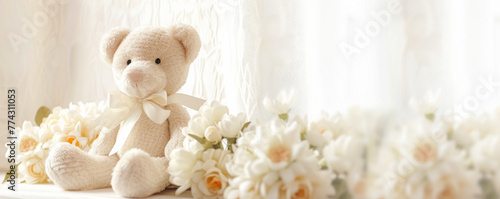 Beige teddy bear with bow sitting beside white flowers on bright background. Soft light nursery decor concept for design and greeting cards. Gentle indoor still life with copy space