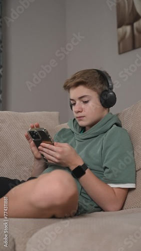 Teenage Boy sitting on sofa using smartphone. Scrolling social media, surfing internet, enjoying time at home alone. Child laying on coach in living room with phone. Vertical shot