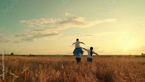 Mom, dad, boy child walk together, family of farmers with child on their shoulders walks through wheat field. Mother, father, little son play, enjoy nature outdoors, kid dream of flying, super hero