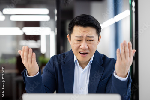 An Asian businessman looking frustrated and confused at his computer in a modern office setting.