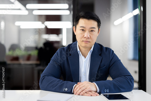 Portrait of a confident Asian businessman seated at a table in a well-lit, contemporary office environment, looking at the camera with focus.