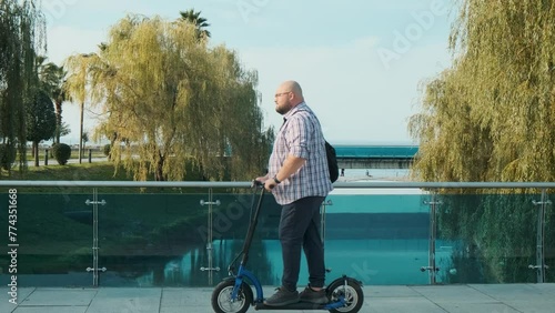 Side view of overweight man riding e-scooter in the city on the btidge. Plus size male driving electric scooter outdoors. Electric and ecological transport. Eco-friendly transportation  photo