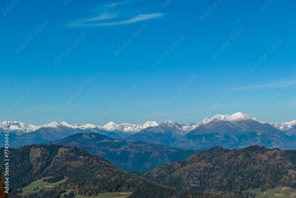 Panoramic view of snow capped mountain ridges of Woelzer Tauern seen from Grebenzen, Gurktal Alps, Styria, Austria. Calm serene atmosphere in Austrian Alps. Idyllic forest in foreground. Wanderlust
