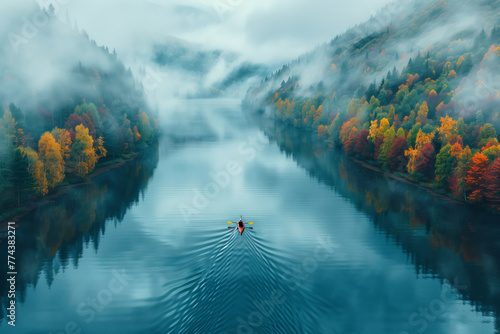 An aerial view of a man paddling a kayak down a river surrounded by autumn trees. The water is calm, the sky is cloudy, and there is morning fog. Peaceful, with the man enjoying the beauty of nature.