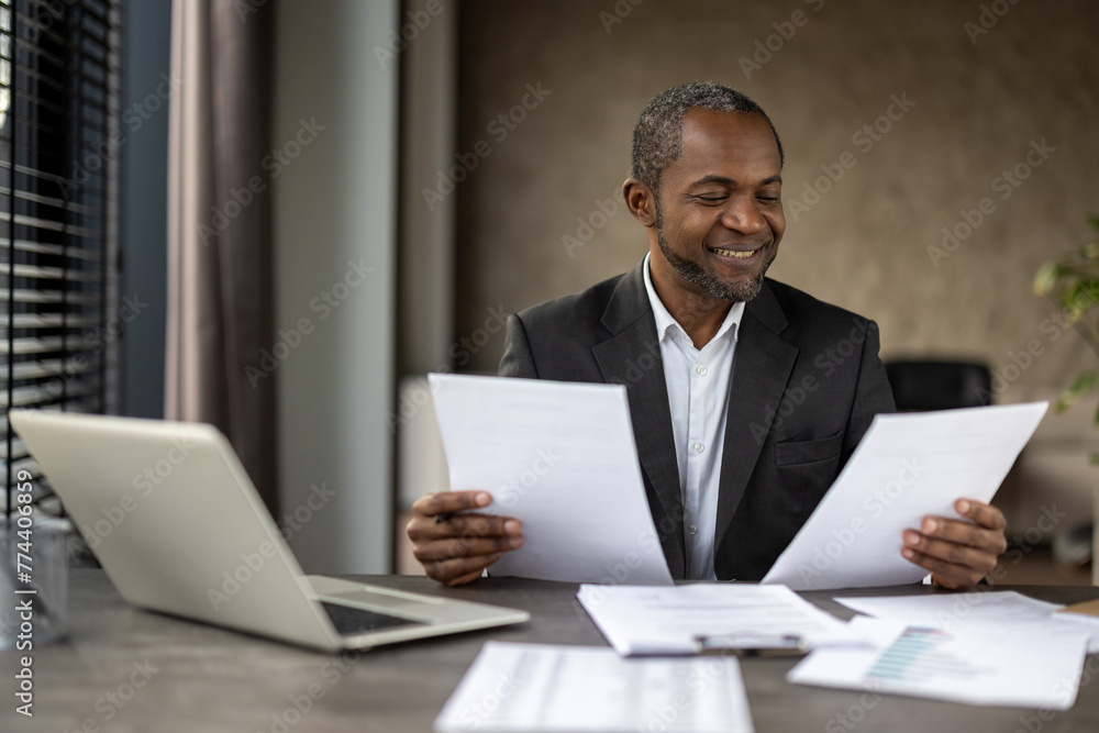 A professional in a suit carefully analyzes documents, surrounded by a laptop and reports on a desk in a well-lit office. A professional in a suit carefully analyzes documents, surrounded by a laptop and reports on a desk in a well-lit office.