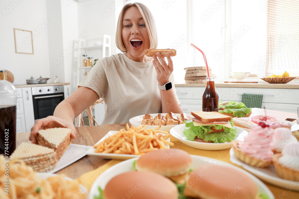 Beautiful woman at table full of unhealthy food in kitchen. Overeating concept