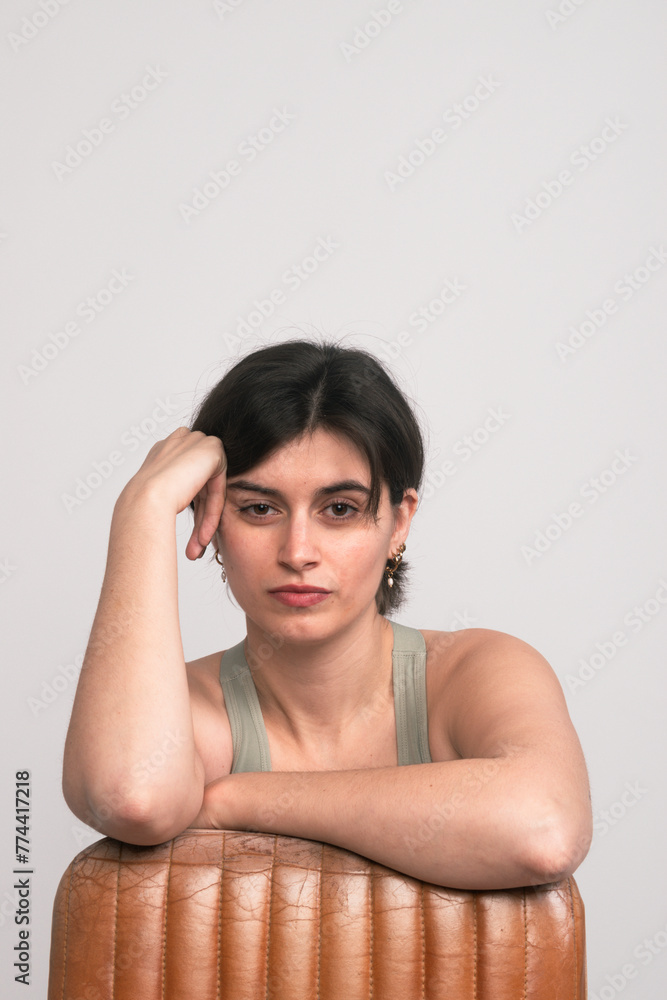 A young woman casually supports her head with her hand while leaning against a brown leather chair, gazing directly at the camera. A young woman casually supports her head with her hand while leaning against a brown leather chair, gazing directly at the camera.