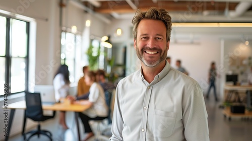 A smiling CEO standing in a bright, open-concept workspace with colleagues working in the background