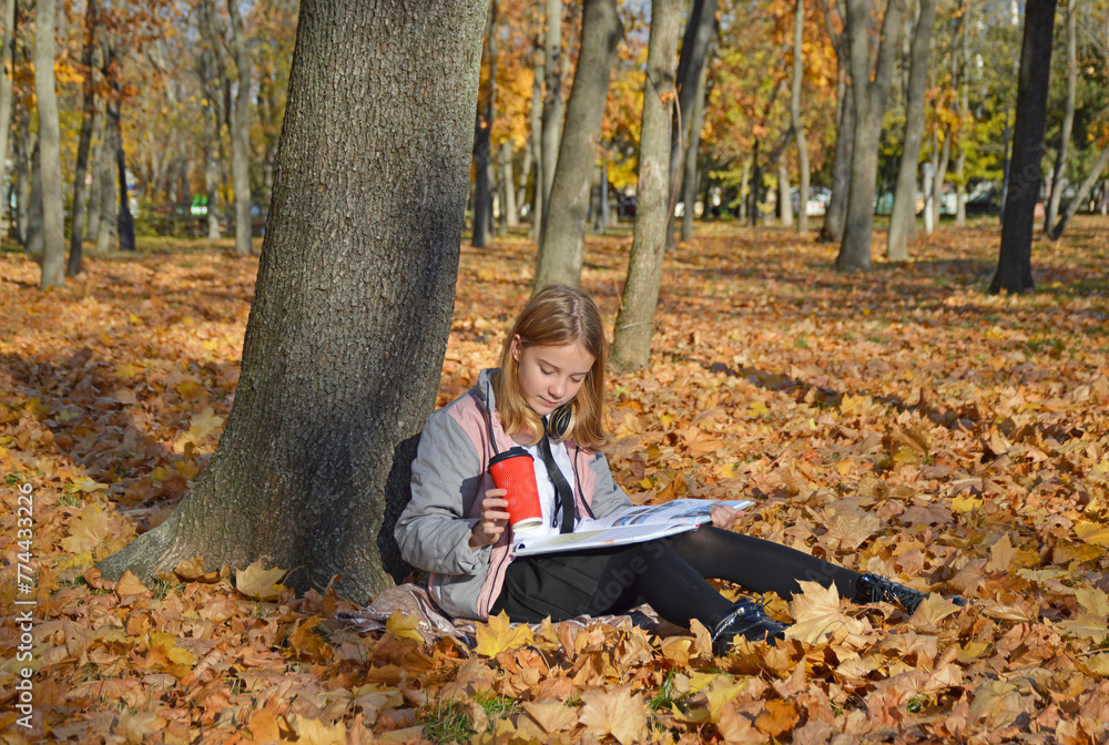 a beautiful girl sits on fallen leaves leaning against a tree trunk in an autumn park. holding a book and a cup of coffee or tea. woman enjoying the warm rays of the sun, hobbies and relaxation a beautiful girl sits on fallen leaves leaning against a tree trunk in an autumn park. holding a book and a cup of coffee or tea. woman enjoying the warm rays of the sun, hobbies and relaxation