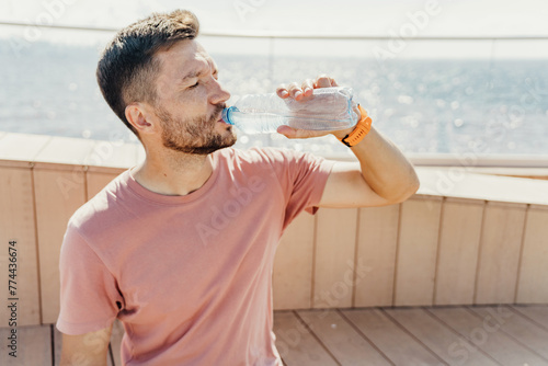 An active man hydrates, drinking water post-exercise on a sunny boardwalk, with the tranquil sea in the background, highlighting the essence of a healthy lifestyle.