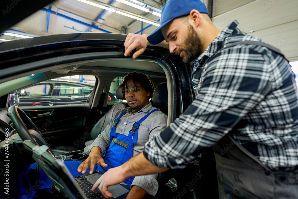 Mechanic colleagues connected the car to diagnostics. One in the driver's seat, the other outside, discussing findings on the computer screen, working together to identify the issue. Mechanic colleagues connected the car to diagnostics. One in the driver's seat, the other outside, discussing findings on the computer screen, working together to identify the issue.
