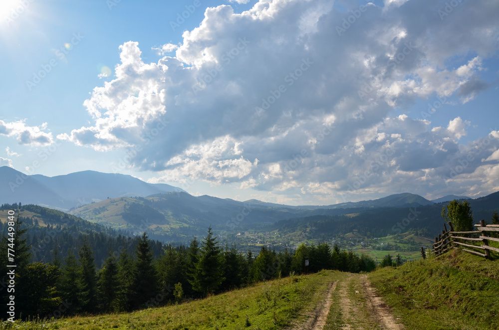 A well-trodden path leads into a valley of the Verkhovyna village nestled amidst rolling hills, adorned with lush greenery under the sky is painted with clouds filtering the golden sunlight