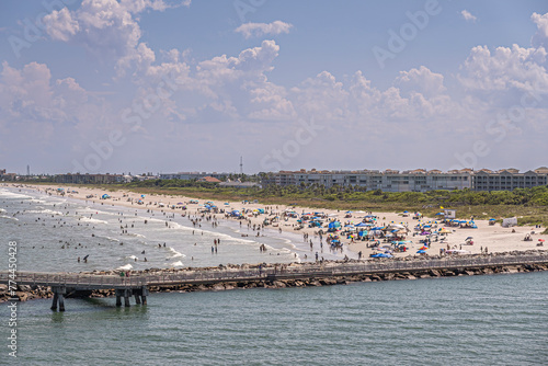 Port Canaveral, Florida, USA - July 30, 2023: Long sandy beach, full of bathers in water and on sand, stretches out as far as one can see, south  of Fisherman Pier. Buildings behind dunes