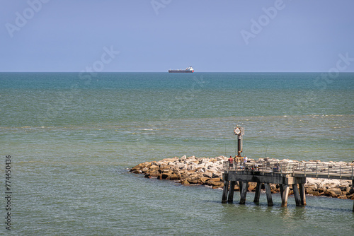 Port Canaveral, Florida, USA - July 30, 2023: Tip of Fisherman pier with some real fishers surrounded by Atlantic Ocean waters under blue sky. Large tanker vessel on horizon