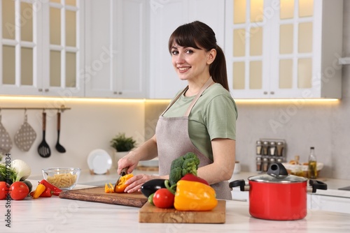 Happy young housewife cutting bell pepper at white marble table in kitchen