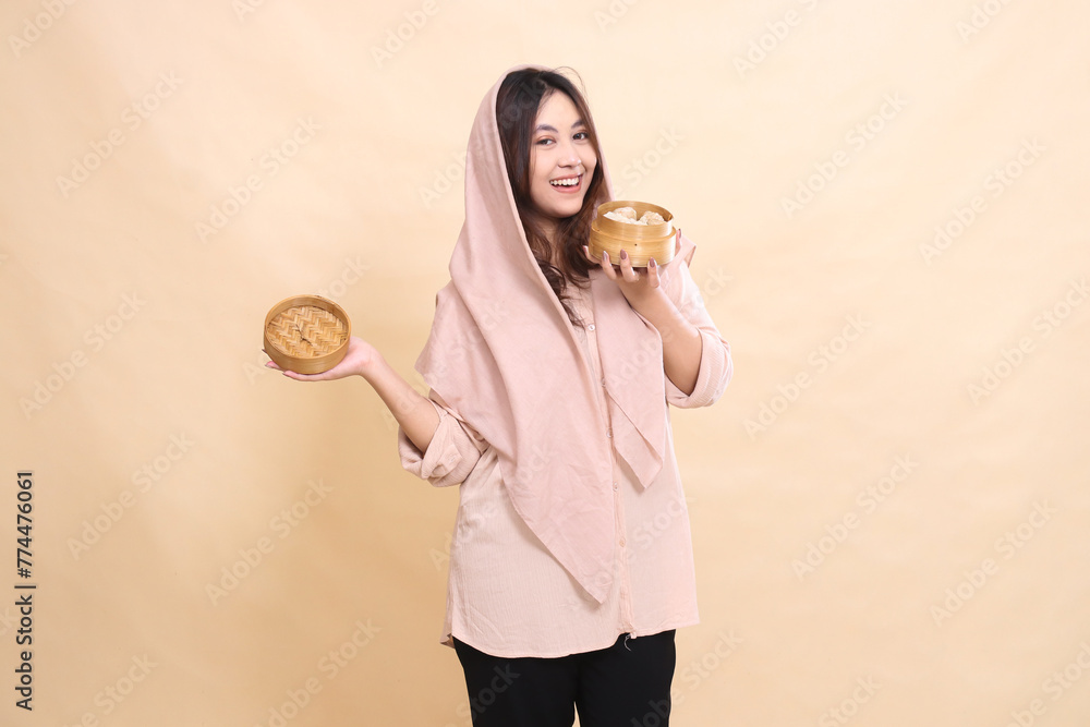 Happy Asian Muslim woman looking at the camera with her hand carrying a wooden bowl sideways containing dimsum. Happy Asian Muslim woman looking at the camera with her hand carrying a wooden bowl sideways containing dimsum.