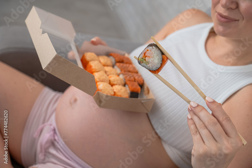 A pregnant woman sits on the sofa and eats rolls from a box. Food delivery. 