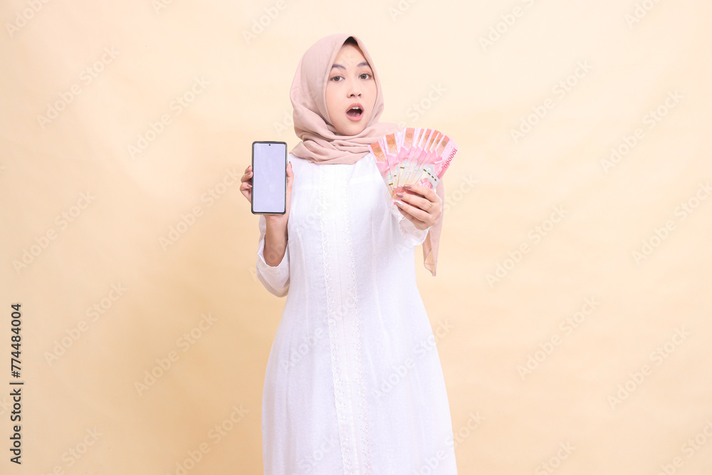 woman indonesia young muslim wearing a hijab is shocked holding a cellphone gadget and rupiah money in her hand. Lifestyle, business and commercial concept woman indonesia young muslim wearing a hijab is shocked holding a cellphone gadget and rupiah money in her hand. Lifestyle, business and commercial concept