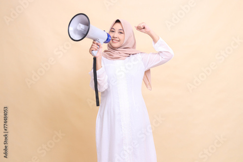Asian Muslim woman wearing a hijab tilted to the right of the camera, smiling, holding a loudspeaker megaphone and clenching her fists upwards. Technology, broadcast and promotion concept
