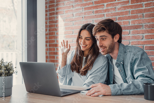 Happy young couple laughing watching funny video or comedy movie online, cheerful man and woman having fun enjoying videocall looking at laptop screen and smiling sitting on sofa at home together. photo
