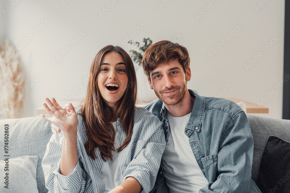 Head shot positive loving bonding married couple sitting on comfortable sofa, looking at camera, holding video call with friends. Happy family bloggers recording video for personal channel at home..