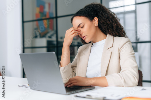 Overworked brazilian or hispanic curly business woman, company employee, working on a laptop in modern office, has a headache, migraine, massages nosebridge with eyes closed, feels stressed and tired