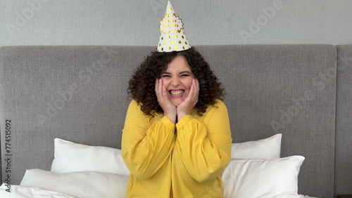 Horizontal 4K video. Beautiful young white brunette childless woman with curly hair wearing yellow shirt and party hat in hotel room looking at camera, laughing and smiling. Concept of fun, joy, smile photo