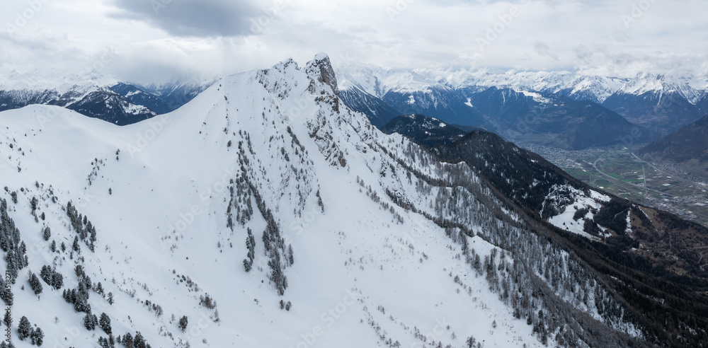 Aerial shot of Verbier, Switzerland's snow clad, rugged mountains, perfect for skiing with steep peaks and valleys. Overcast skies bring drama to the scene, with a town nestled in the mountains.