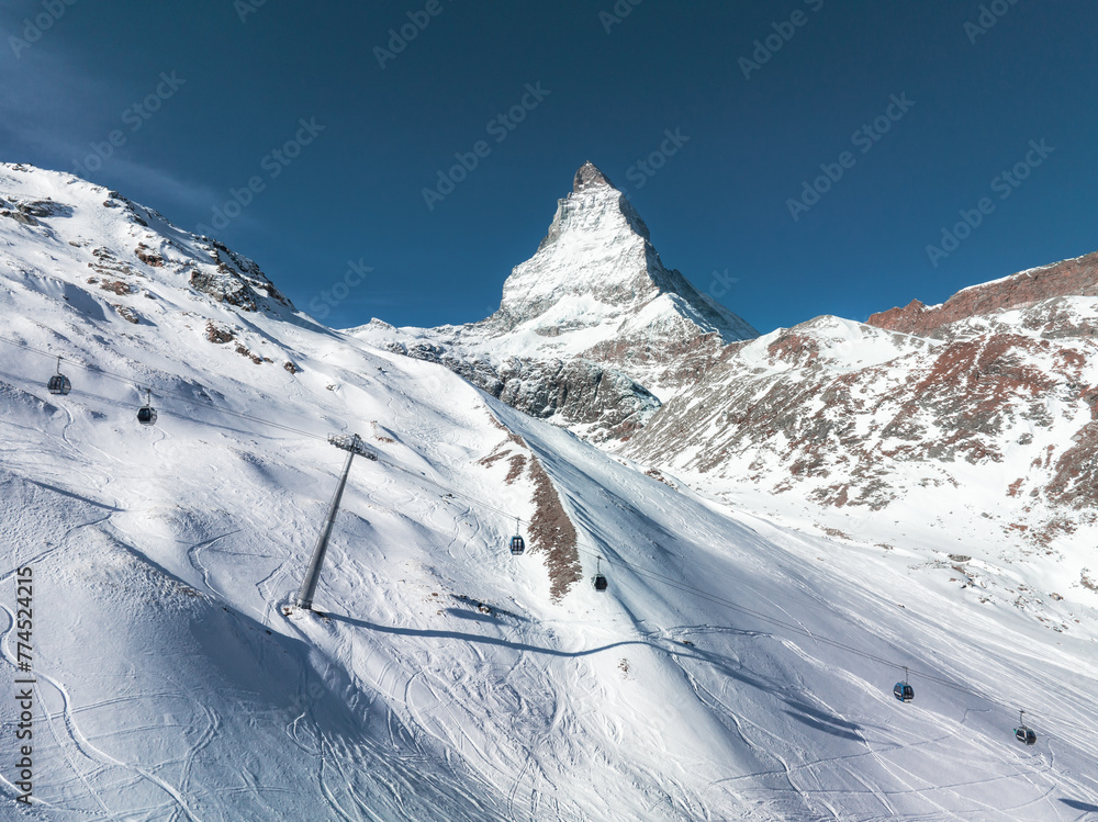 An aerial view of Zermatt, Switzerland, shows the Matterhorn and ski slopes with cable cars. The blue sky and fresh tracks underline its winter sports appeal.
