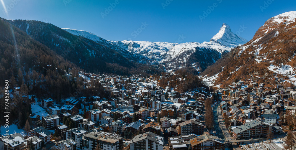 Aerial shot of Zermatt, a tranquil Swiss Alps ski resort, displays chalets and modern facilities under snow. The Matterhorn and snow capped peaks loom behind, creating a serene, majestic winter haven.