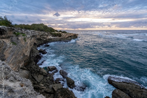 The sun slowly rises over jagged cliffs, which meet the rough turquoise waters of the Pacific Ocean along the Mahaulepu Heritage Trail in Koloa, Hawaii on the island of Kauai.