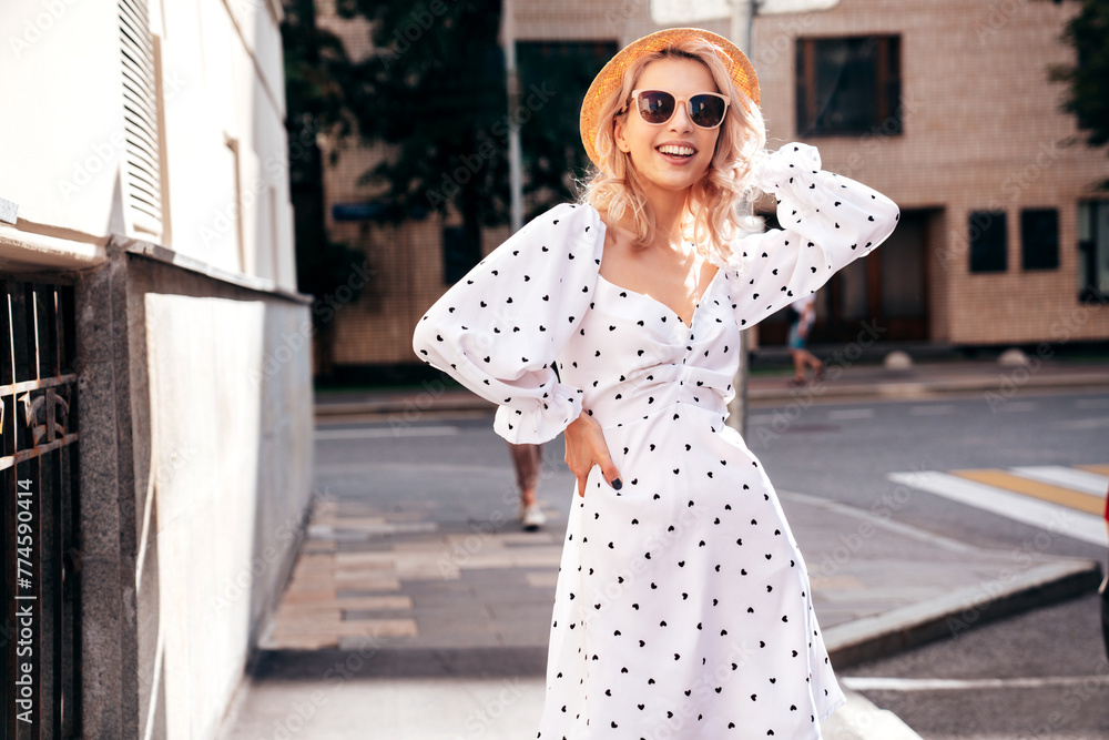 Naklejka premium Young beautiful smiling blond woman in trendy summer white dress. Sexy carefree woman posing in the street at sunset. Positive model outdoors at sunny day. Cheerful and happy. In hat, sunglasses