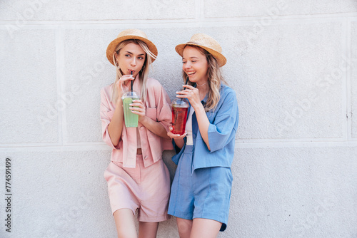 Two young beautiful smiling hipster female in trendy summer clothes. Carefree women posing outdoors. Positive models holding and drinking fresh cocktail smoothie drink in plastic cup with straw
