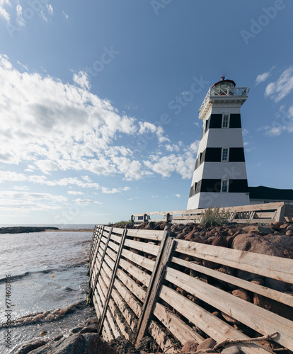 vue sur un phare ligné noir et blanc en bord de mer avec un brise-vague en bois en avant plan