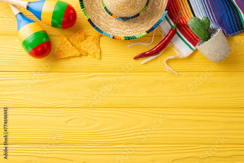 Festive Cinco de Mayo top view setup with sombrero, maracas, and cactus. Vibrant serape, nacho and chili pepper adorn the yellow wooden desk. Top view with space for text