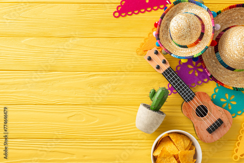 Cinco de Mayo top view scene: sombreros, vihuela, and a cactus on display. Colorful flag garland, and spicy nacho adorn the yellow wooden desk. Space for text