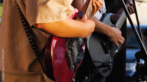 Close-up of a man playing guitar with another guitarist in the background. They are performing together at an outdoor concert. The emphasis is on the interaction between the musician and the guitar.