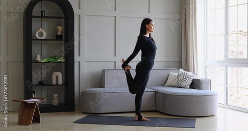 A woman leading a healthy lifestyle performs the Natarajasana exercise at home, king of the dance pose, training in a black sports overalls standing on the mat, full-length view