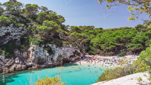 Turquoise shallow clear water, white fine sand beach and rocky cliffs with green pine trees, picturesque landscape of Cala Macarelleta in Menorca Island Spain, crowded with tourists in summer holiday photo