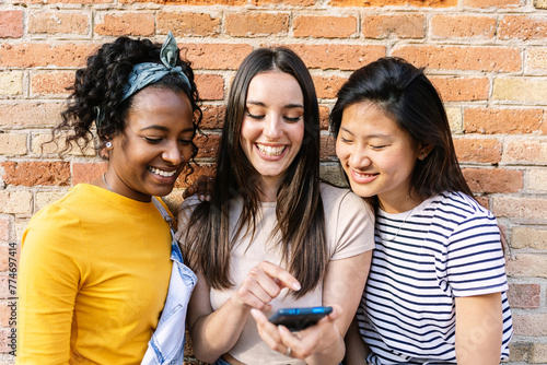 Happy young girls using mobile phone standing over brick wall background outdoors. Social media concept.