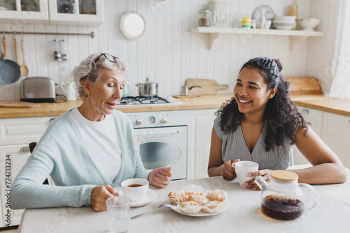 Indoor portrait of two people, black young female volunteer visiting with lonely retired female with gray hair, having nice conversation sitting at kitchen table, drinking tea with sweets