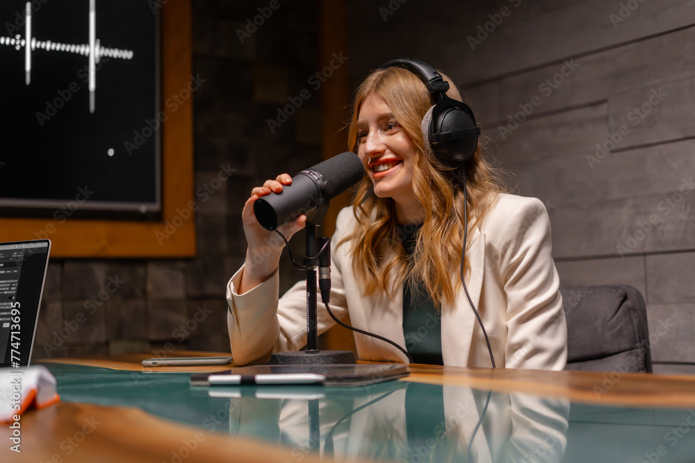 A woman with headphones is chatting into a microphone while sitting at a table. Podcast studio concept - obrazy, fototapety, plakaty 