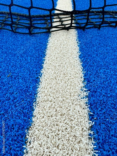 Close-up view of a line and texture of blue paddle tennis court and lonely yellow tennis ball. Artificial grass paddle court surface.