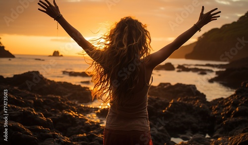 A woman with long hair is standing on a rocky beach, arms raised in the air