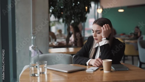 beautiful, sad young woman sits in a cafe in business attire, angrily closes her laptop, tapping on the table with her hand photo