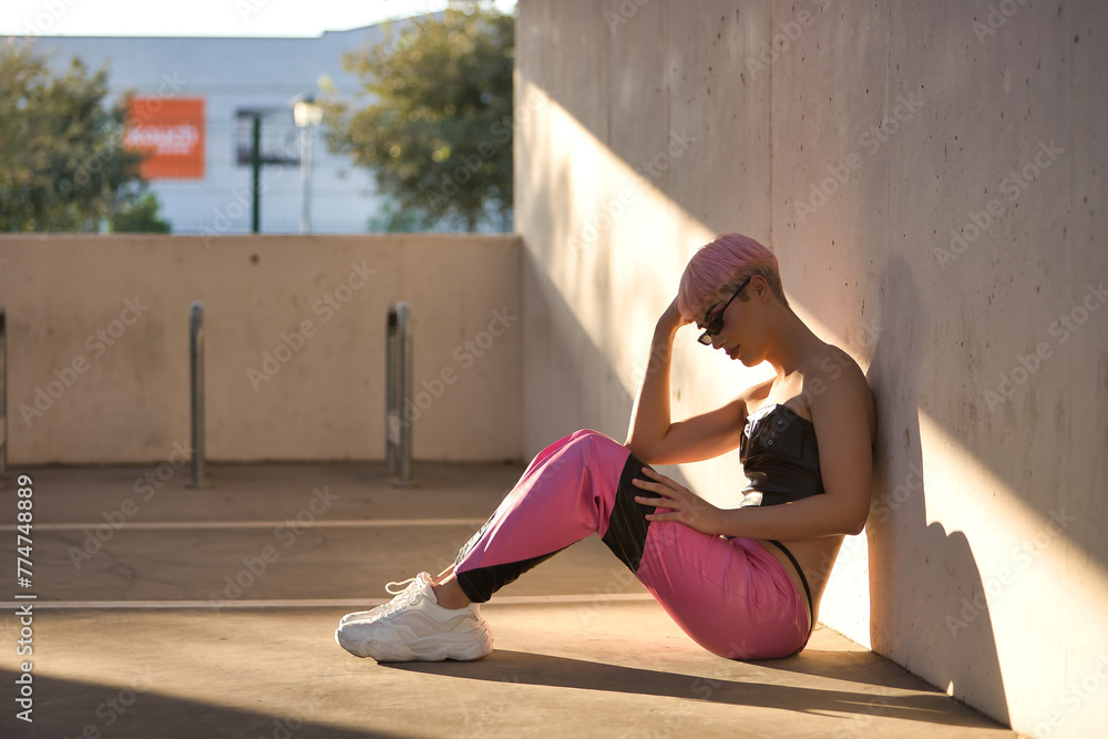 Obraz premium Young attractive gay man, heavily makeup, with pink hair, sunglasses, top and leather pants, alone, depressed and sad, sitting on the ground getting a sunbeam at sunset. LGTBIQ+ concept, gay, pride.