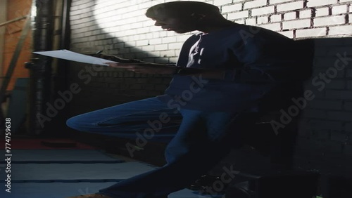 Vertical lowkey shot of young Black male standup comedian leaning on brick wall in dark corridor and reading his monologue once again before going on stage to make comedy performance photo