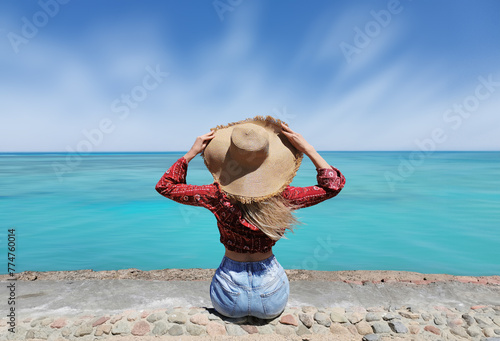 A woman in a red shirt and jeans posing with a big hat with a blue sea view. Fashion minimalism long exposure shot.